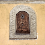 Das gut erhaltene Weinfenster in der Via Torta. An der gelben Mauer, mit Stein umrandet und mit einem gut erhaltenen, braunen Holztürchen versehen.