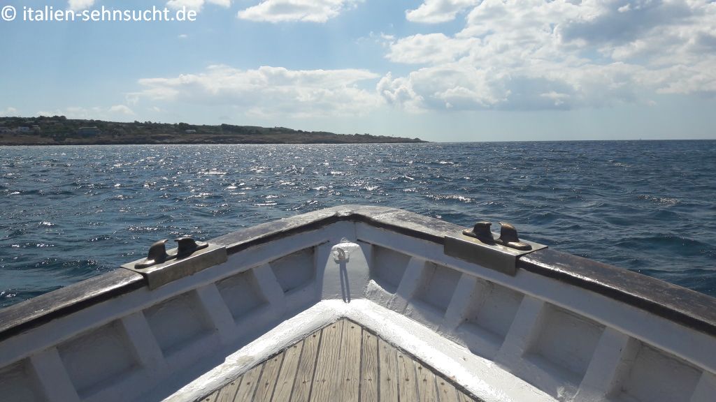 Vom Boot namens Maretta aus sieht man dessen Spitze und blickt auf Meer und Küste. Der Himmel ist hellblau, ein paar Wolken tummeln sich.
