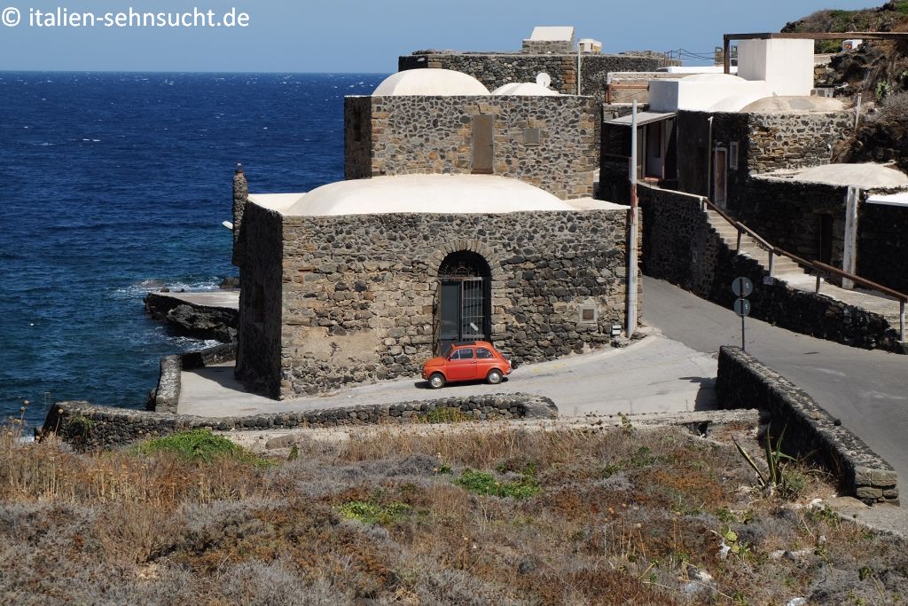 Pantelleria: Ein roter alter Fiat Cinquecento steht vor einem Dammuso, dem charakteristischen Häusertyp für Pantelleria. Direkt dahinter beginnt das blaue Meer.